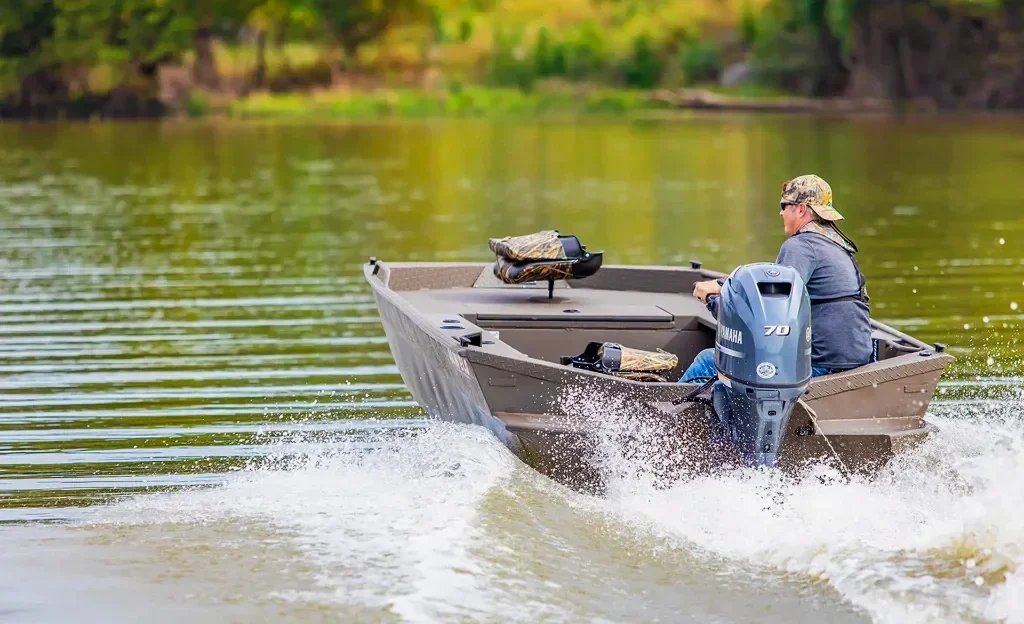 Aluminum fishing boat with a Yamaha 70 outboard underway on a calm lake, with a driver in a camouflage cap at the tiller.