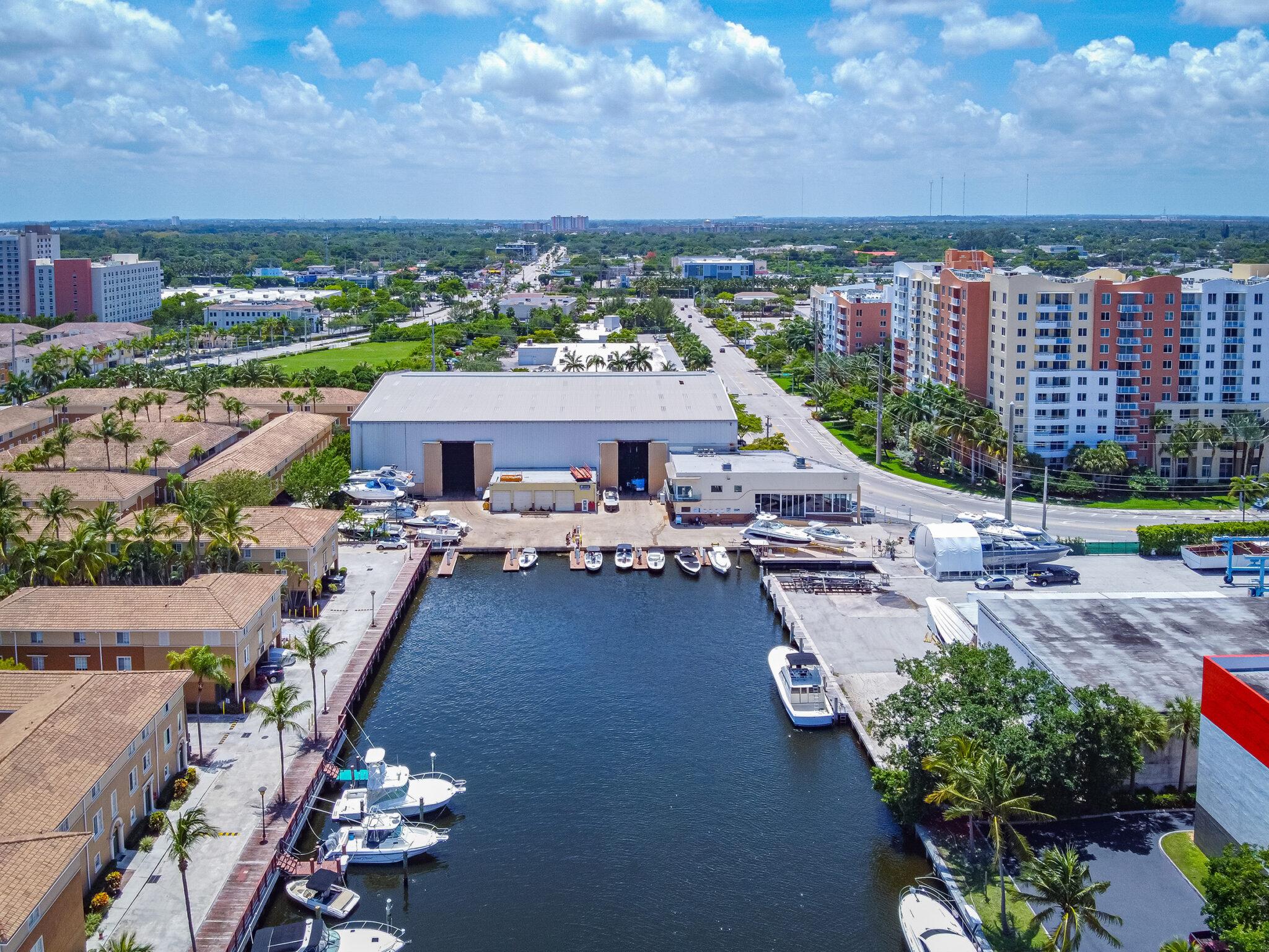 Aerial view of a boatyard with a large boat storage hangar and docked boats along a canal, with condominiums nearby, in South Florida.