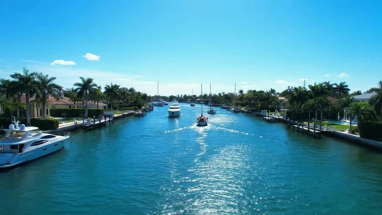 Sunny canal lined with palm trees, waterfront homes, docked yachts, and sailboats underway in a residential waterway.