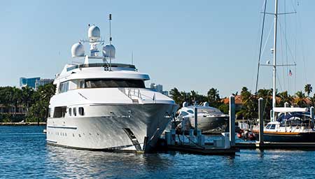 Large white motor yacht moored at a marina dock with sailboats alongside, under a clear blue sky.