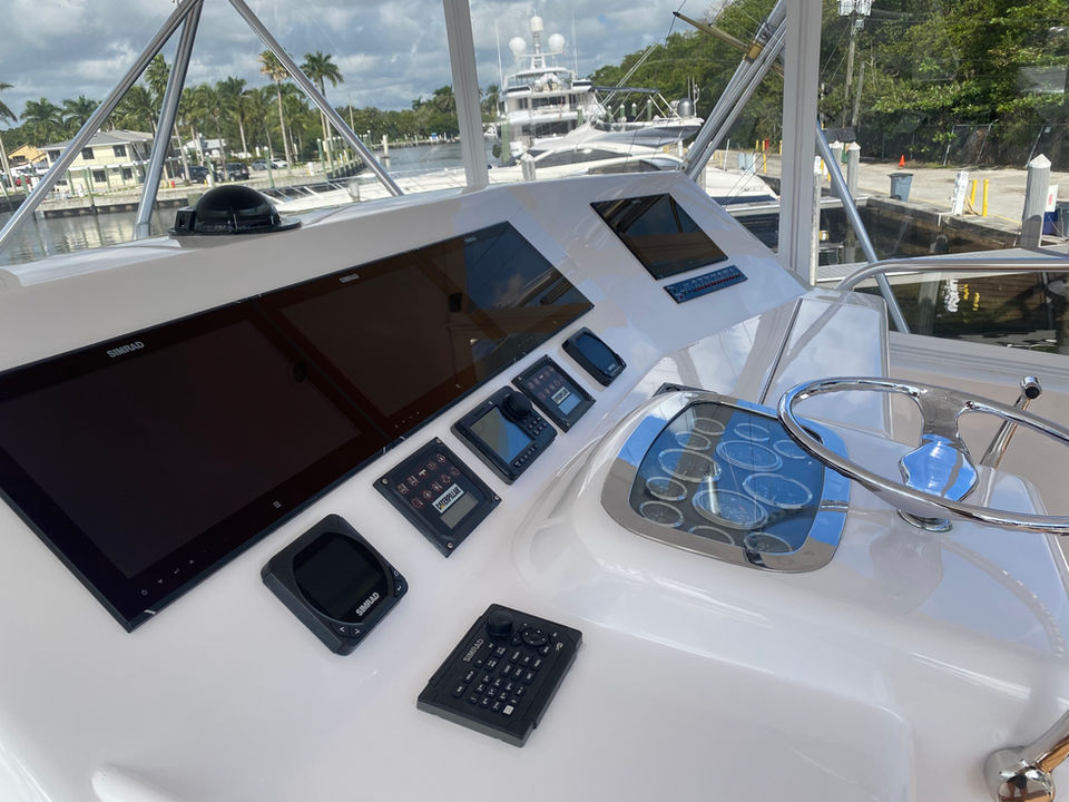 Helm station with Simrad navigation displays and controls on a motor yacht overlooking a marina, with docked yachts and palm-lined waterfront in view.