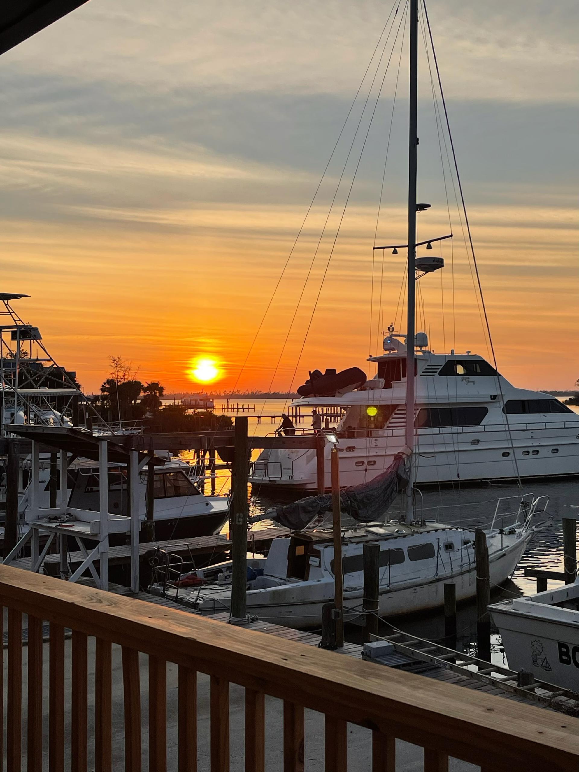 Sailboat and motor yacht docked at a marina with wooden piers at sunset, with the sun setting over the water.