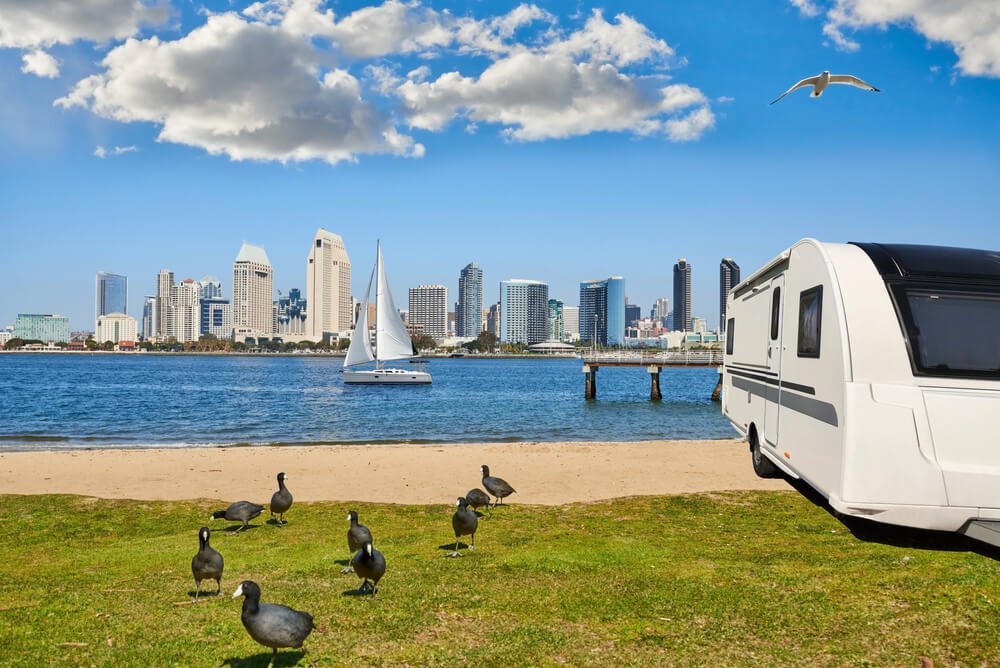Camper trailer parked beside a bay with a sailboat and downtown San Diego skyline in the background.