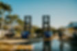 Tall steel boat lift towers over calm water at a marina shoreline lined with trees.