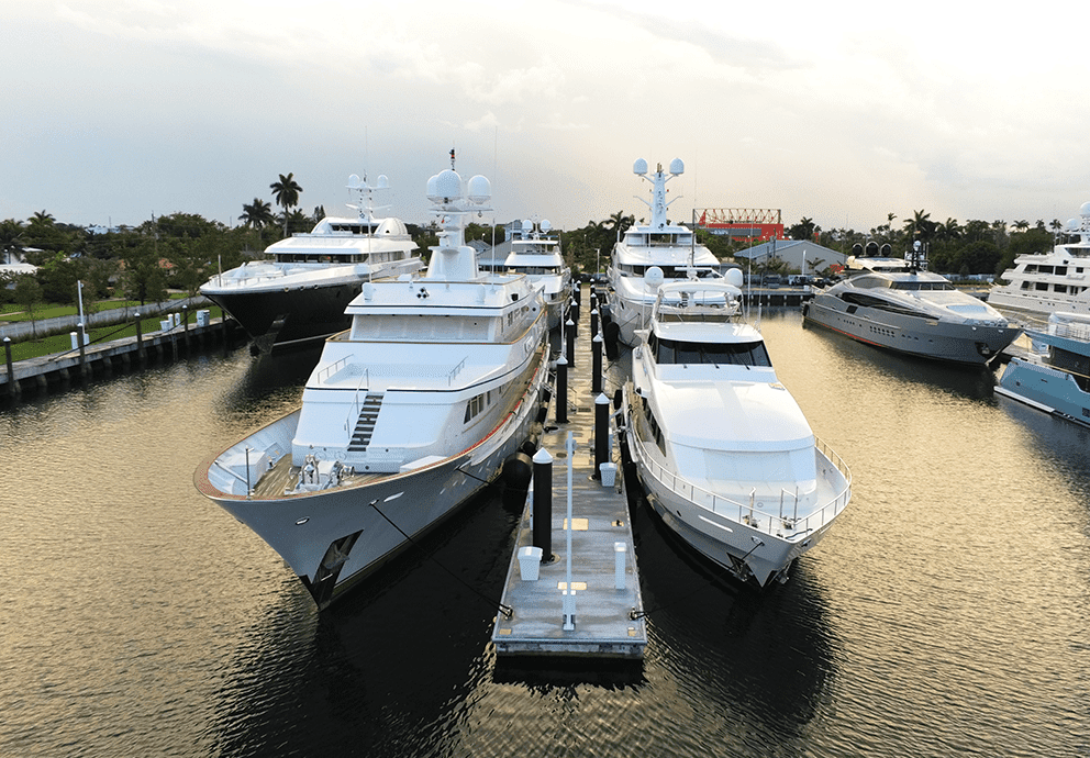 Several large superyachts moored along a canal dock in a waterfront marina, with palm trees lining the shore.