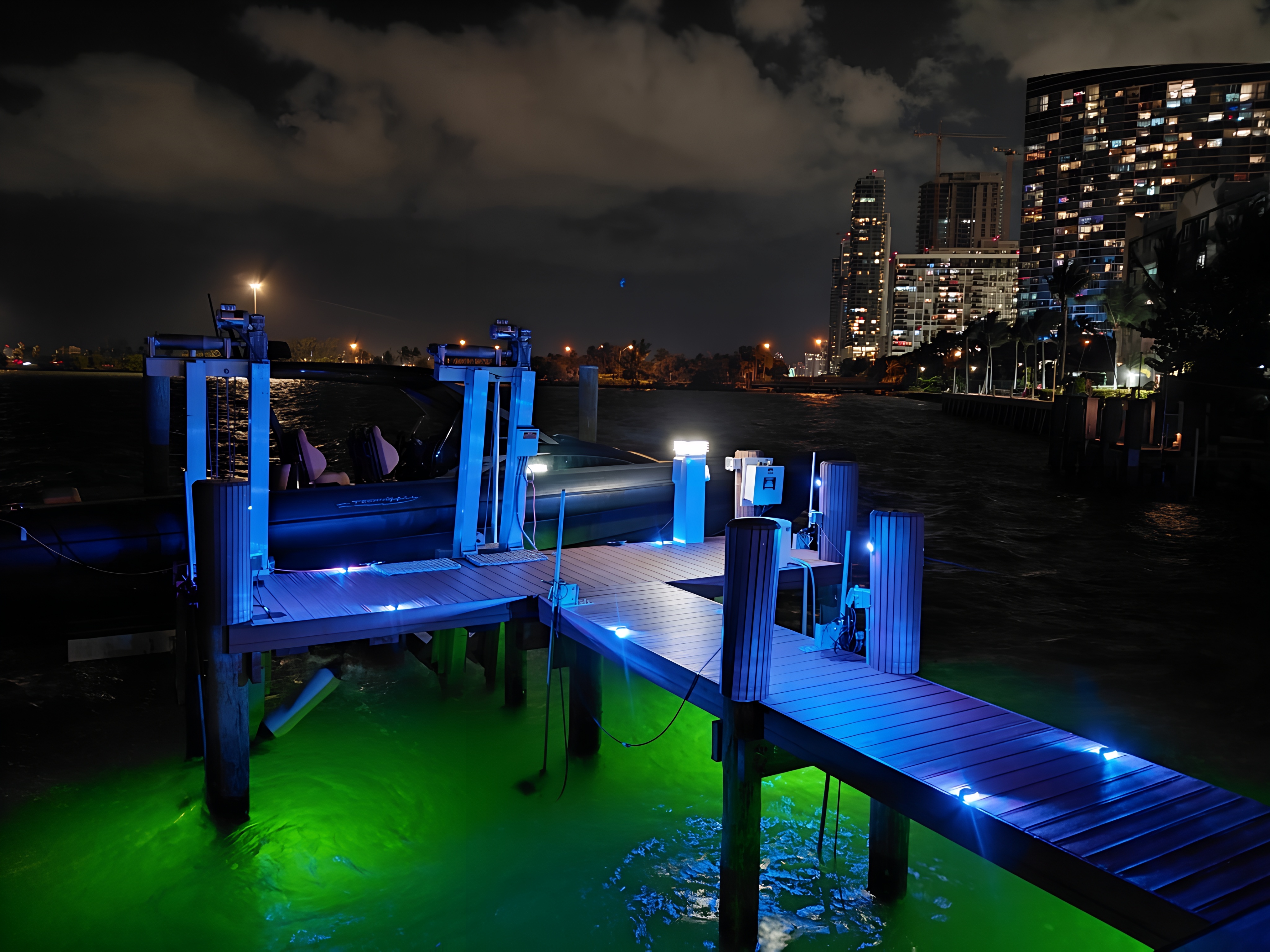 Illuminated boat lift and dock with LED deck and underwater lighting beside a moored boat, with a city skyline at night, business and town not identified.
