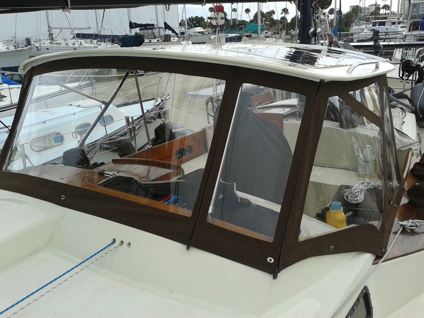 Clear vinyl dodger and cabin enclosure with dark wood trim on a cream-colored sailboat at a marina dock.