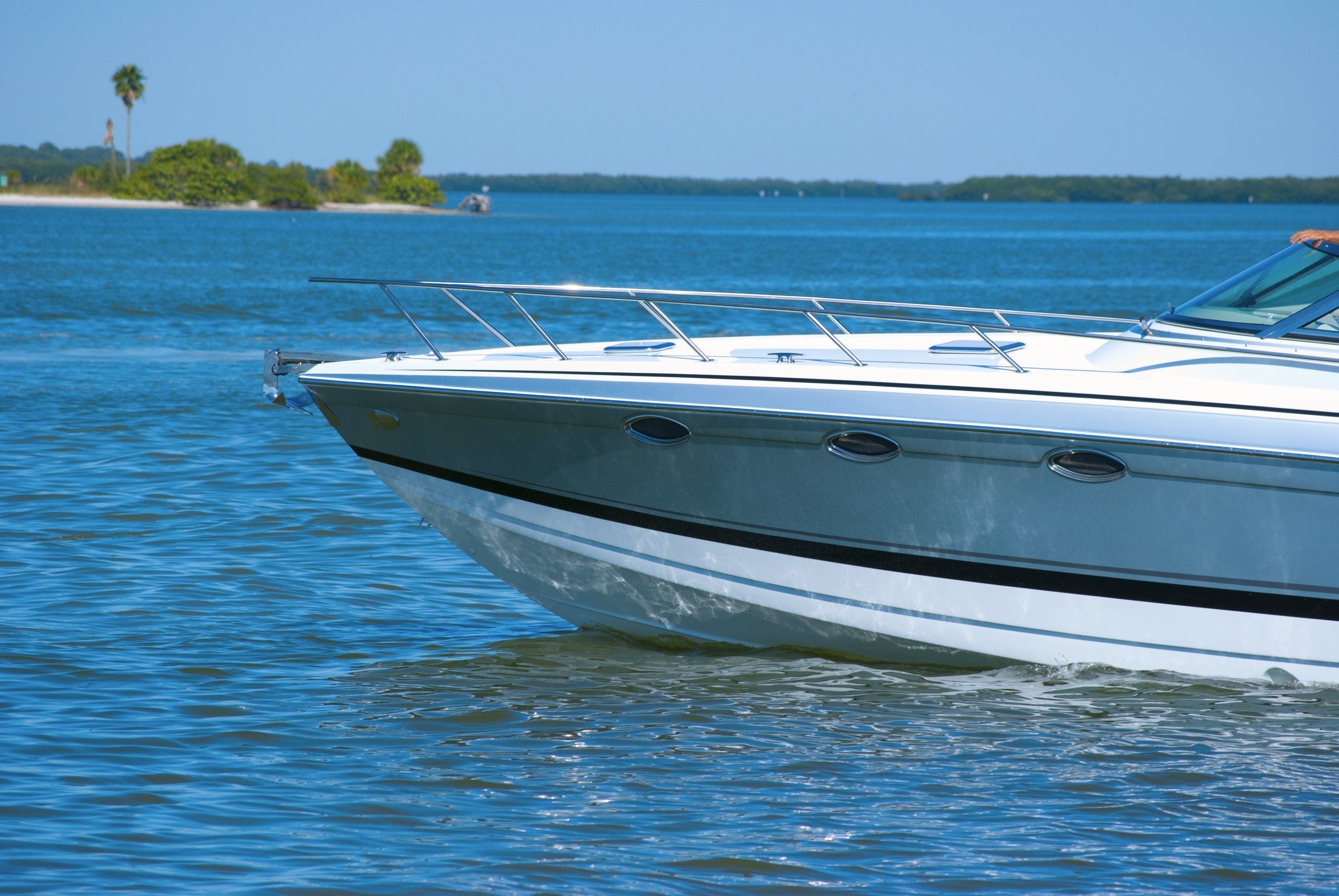 Bow of a white and gray cruiser boat with stainless steel railings moving across open water toward a tree-lined island.