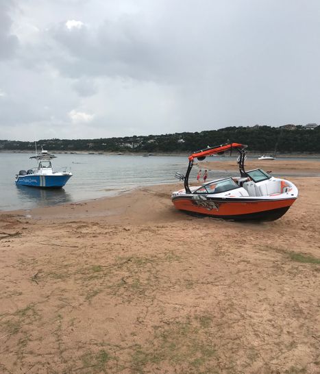 Orange and white wake boat beached on a sandy shoreline next to a blue and white boat on a lake with wooded hills behind.