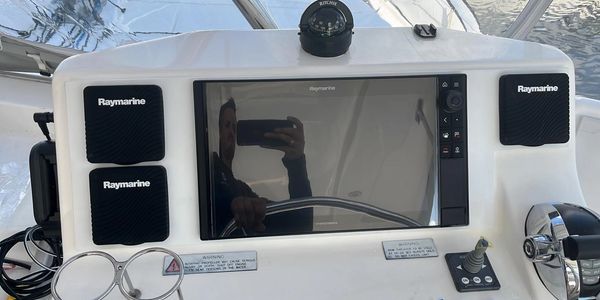 Boat helm station with a Raymarine chartplotter display and Raymarine speakers mounted on the console, with a person's reflection visible in the screen.