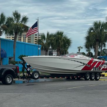 Red and white performance boat on a dual-axle trailer hitched to a black pickup truck in a parking area with palm trees and an American flag, location not confirmed.