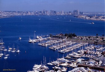 Aerial view of a crowded marina with docked sailboats and yachts, with a city skyline across the bay.