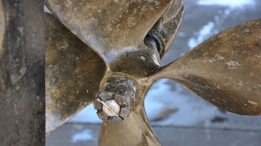 Weathered bronze four-blade ship propeller with a locking nut and cotter pin on the shaft hub, shown in close-up against a blurred background.