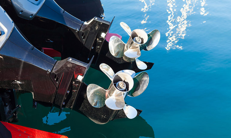Twin outboard motors with polished stainless steel propellers mounted on a boat transom above clear blue water.