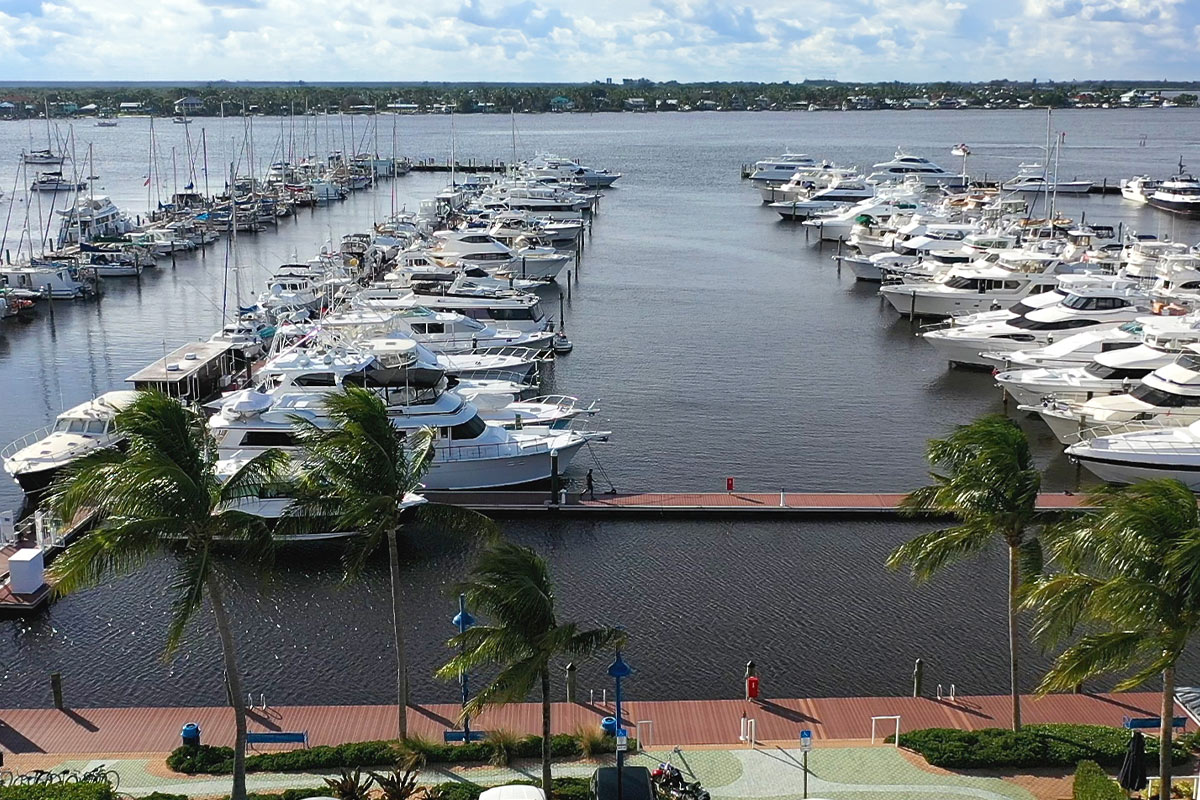 Aerial view of yachts and sailboats docked at a marina with a palm-lined waterfront promenade, town name not visible.