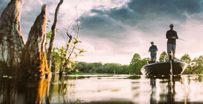 Two anglers fishing from a bass boat on a calm lake lined with cypress trees under a cloudy evening sky.