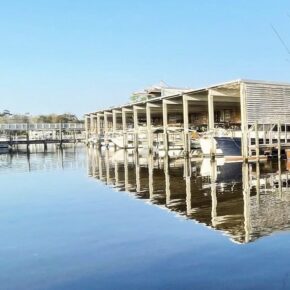 Covered boat slips with white boats moored at a marina dock, reflected in calm water.