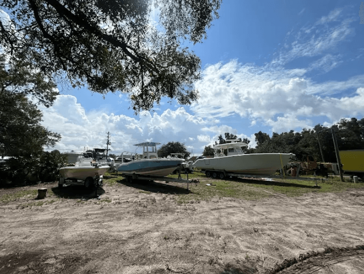 Boats on trailers and cradles in an outdoor storage yard under shade trees, with no business name or town visible.