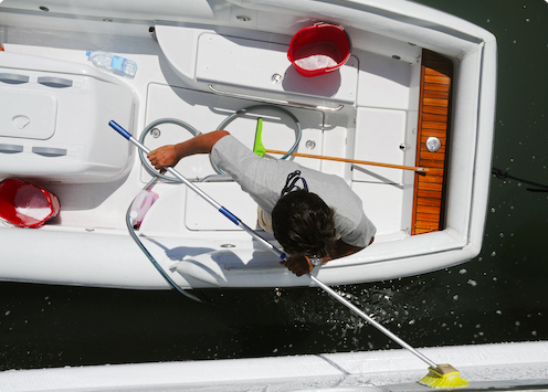 A person scrubbing the deck of a white boat with a long-handled brush while water sprays from the bow area.