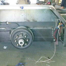 Trailer axle with drum brake and wheel hub on a metal trailer frame in a fabrication shop.