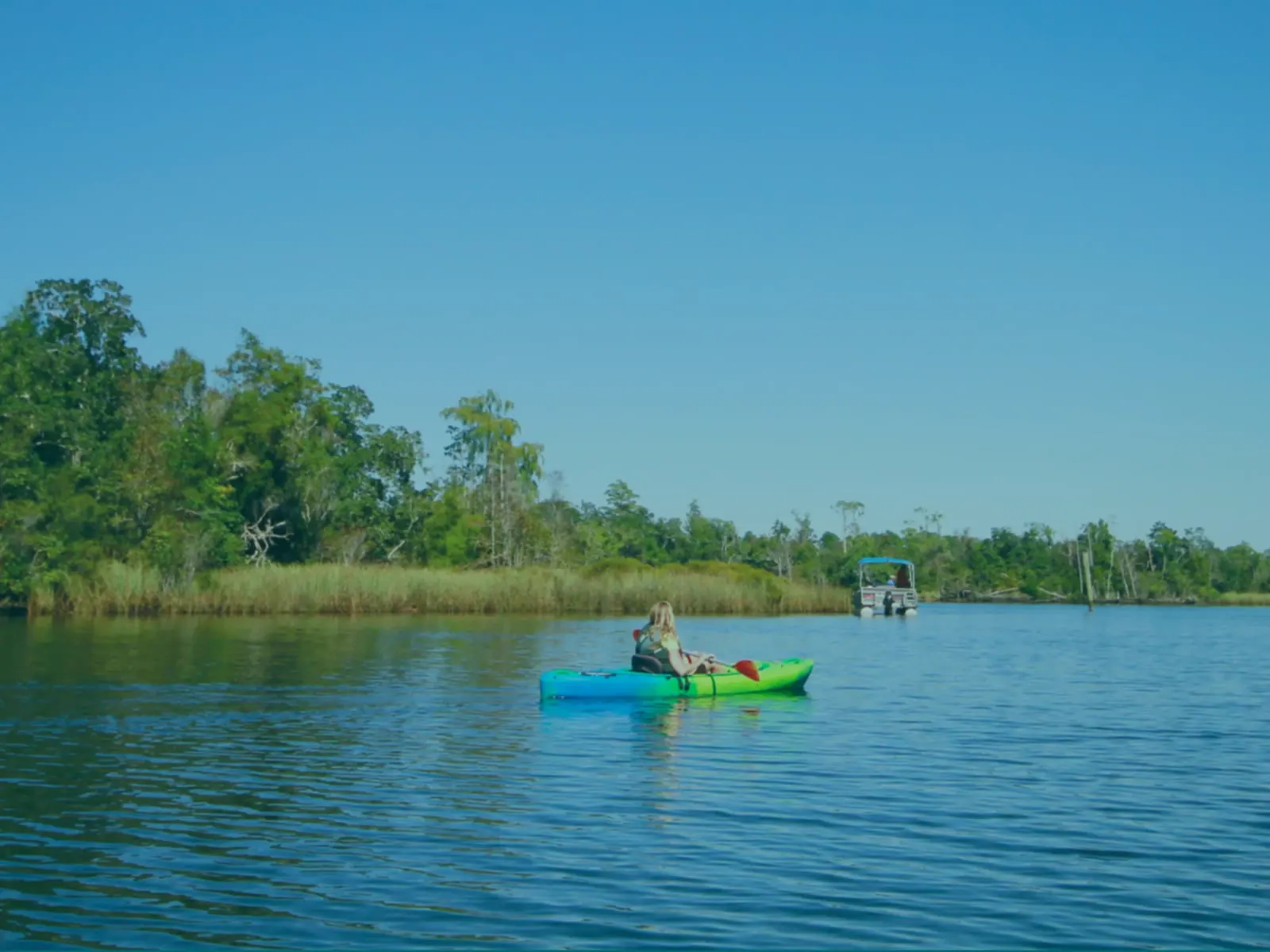 Kayaker paddling a green kayak on a calm tidal river lined with marsh grass and cypress trees, with a pontoon boat behind.