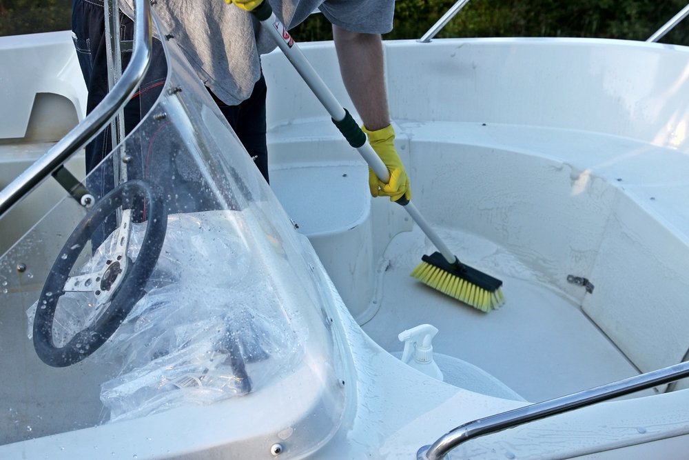 Person scrubbing the white deck and helm area of a boat with a long-handled brush and yellow gloves.