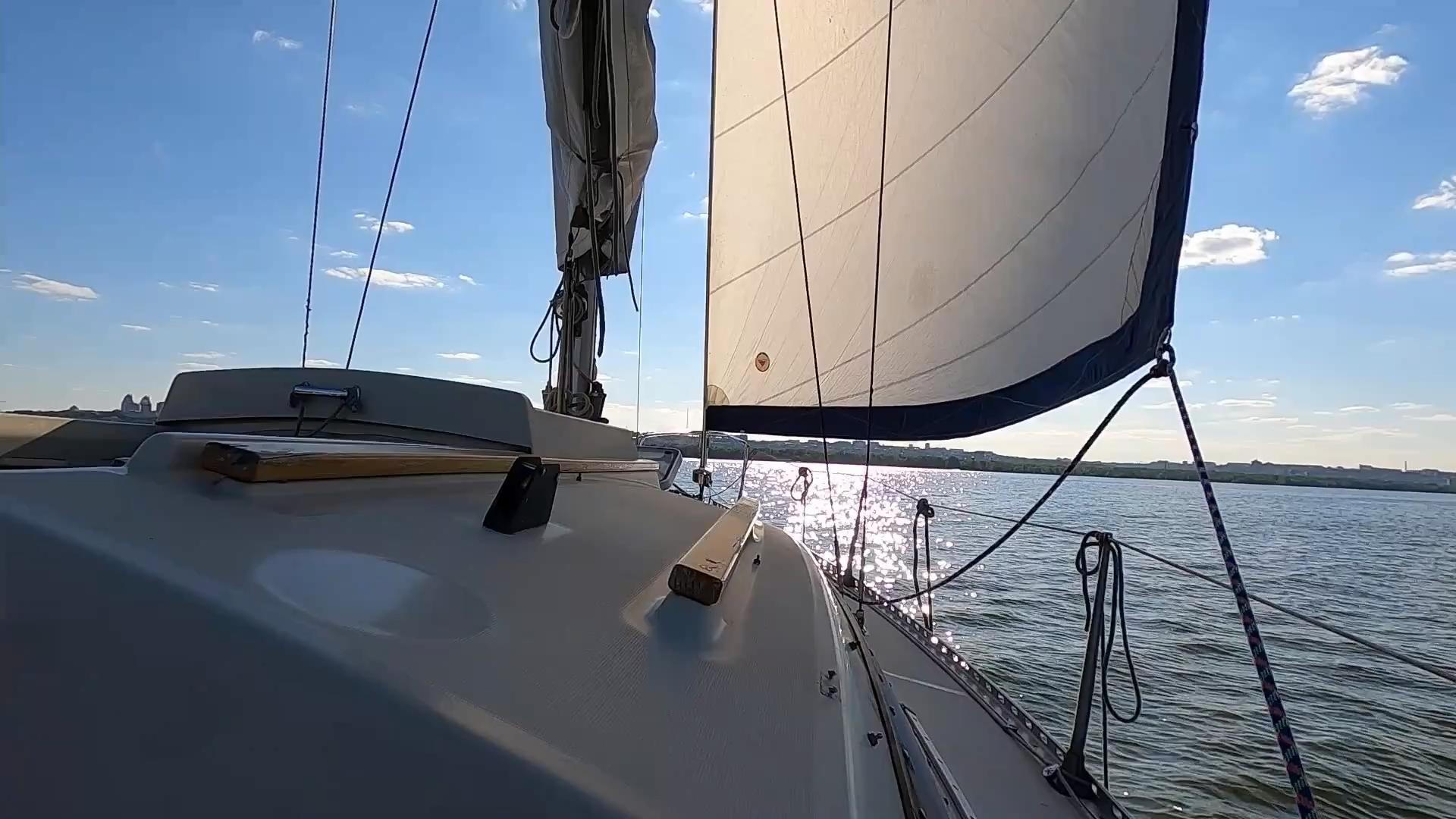 Sailboat under sail on open water with sunlit ripples and a distant city shoreline, viewed from the foredeck.