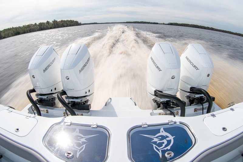 Center-console boat with four white Mercury outboard engines churning a wake across a wide waterway, viewed from the bow.