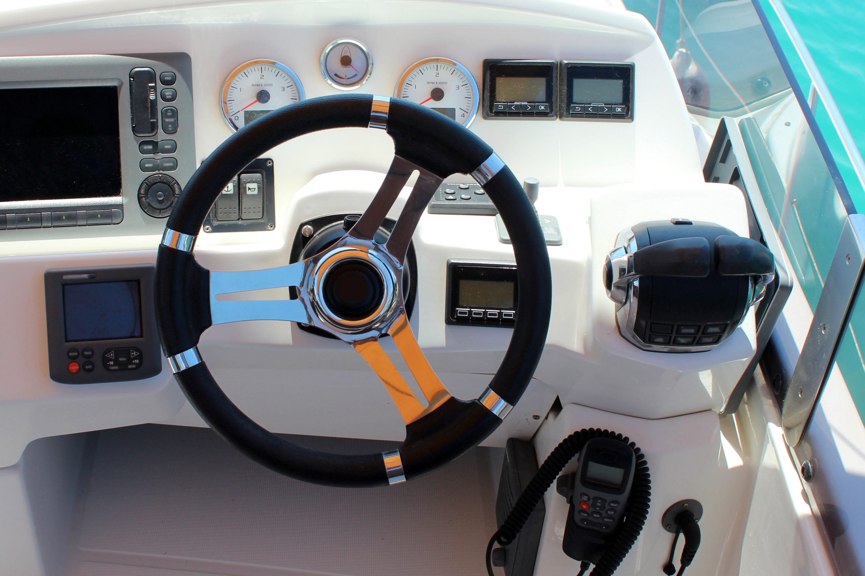 Boat helm station with a chrome-spoked steering wheel, twin tachometers, a chartplotter, and a VHF radio microphone.