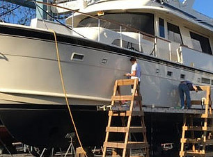 Motor yacht hull being worked on by two technicians on wooden stepladders in a boatyard.