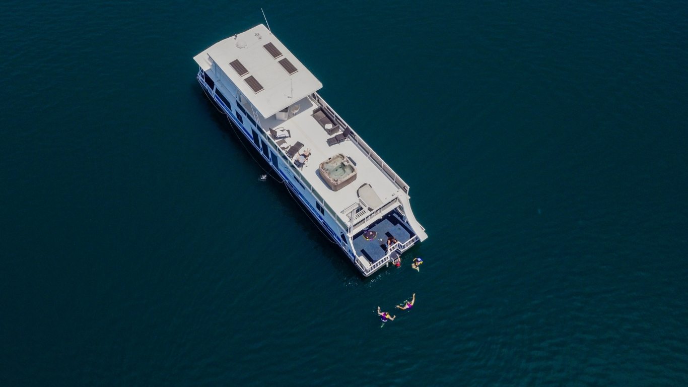 Aerial view of a white houseboat with a hot tub and sun deck anchored on open water, with swimmers beside its stern.