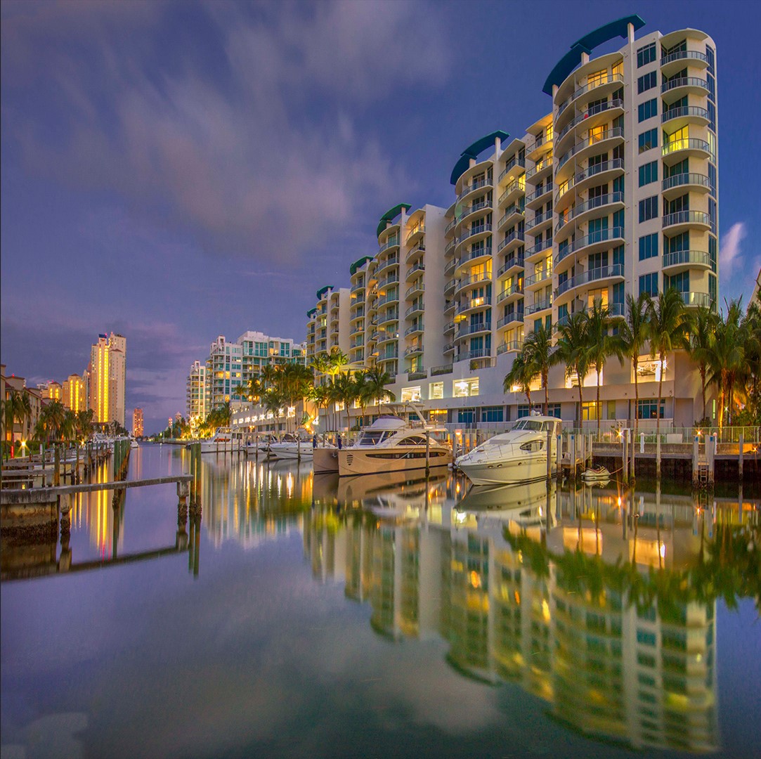 Luxury waterfront condominium towers with yachts docked along a canal at dusk, with palm trees and reflections in the water.