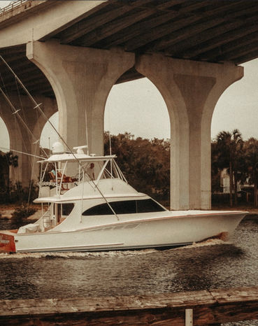 White sport fishing boat with outriggers passing beneath a concrete bridge on a waterway.