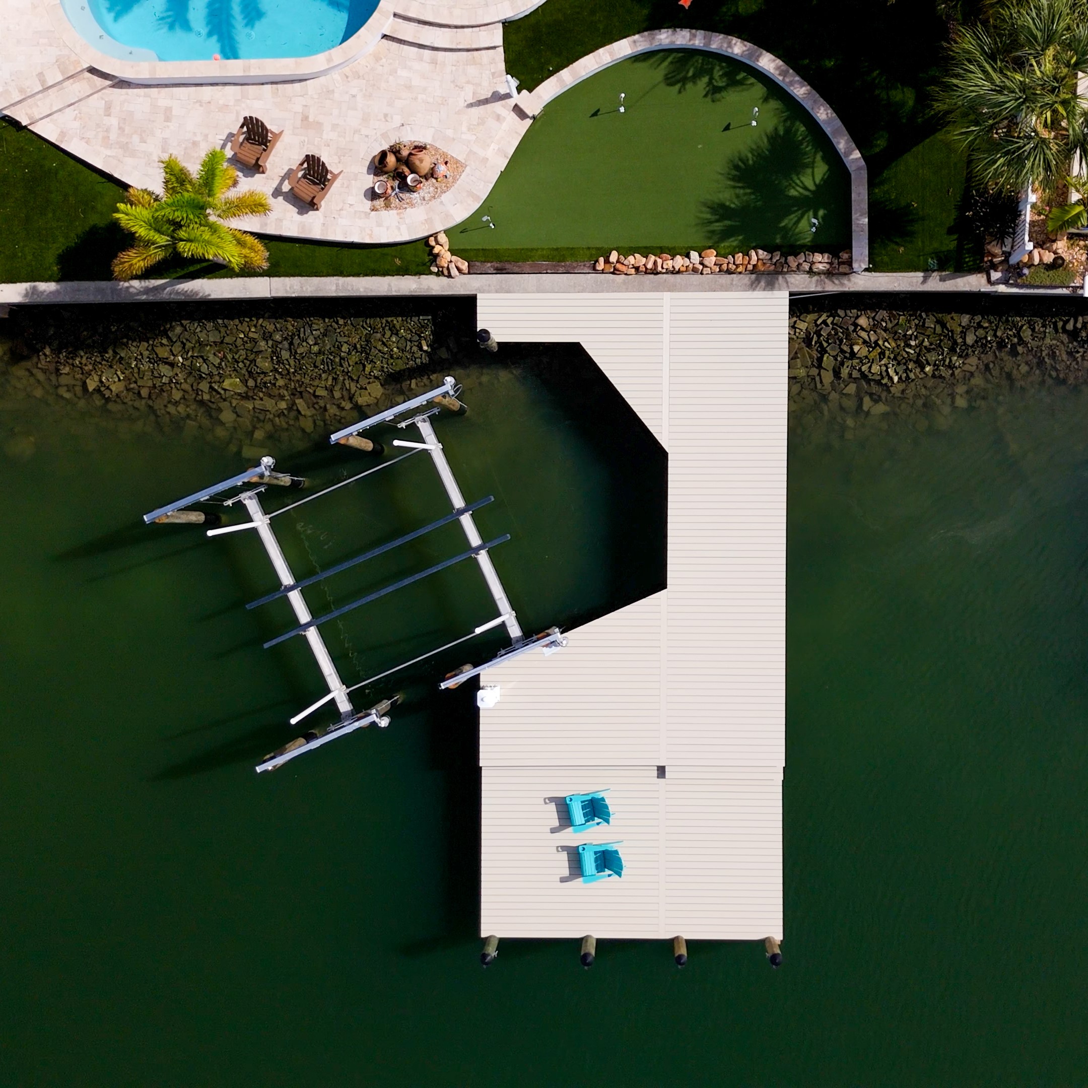 Aerial view of a private waterfront dock with a multi-slip boat lift and white composite decking behind a backyard pool and putting green.