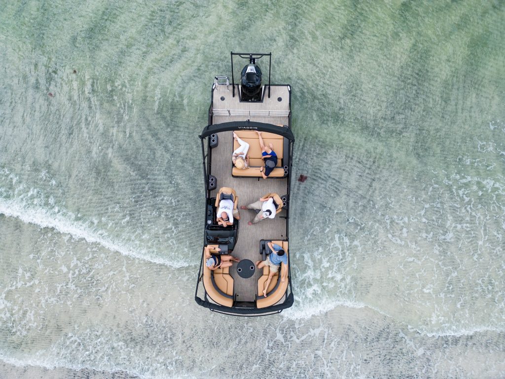 Aerial view of a Viaggio pontoon boat with passengers relaxing on tan seating in shallow turquoise water.