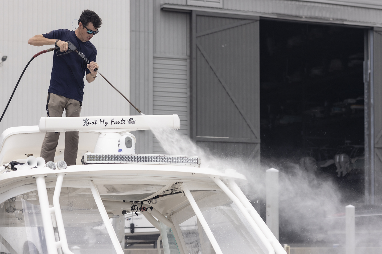 Man pressure washing the white hardtop of a center-console boat named Knot My Fault in front of a boat storage building.