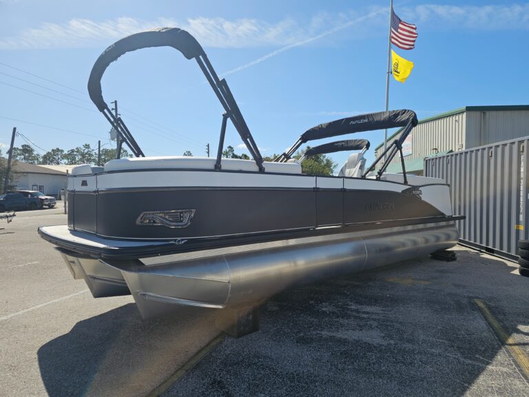 Pontoon boat with a black hull and bimini tops displayed on a dealer lot beside an American flag, in a sunny outdoor setting.