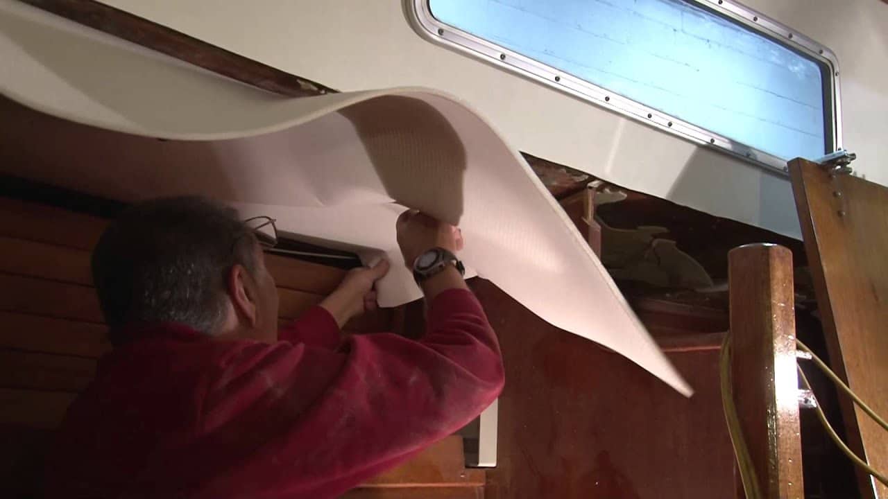 Man installing a white headliner panel in a boat cabin beneath a porthole.
