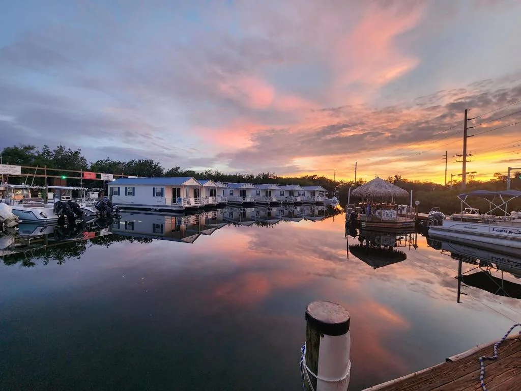 Houseboat row and moored boats at a marina at sunset, with a thatched tiki gazebo dock in view.