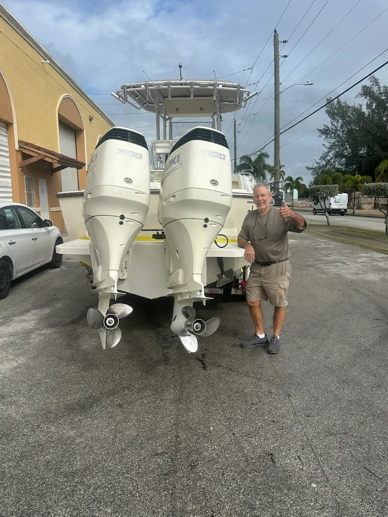 Center console boat with twin Suzuki 300 outboard motors in a paved lot beside a yellow stucco building, with a customer giving a thumbs-up.