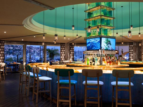 Curved bar interior with pendant lights and windows overlooking a harbor marina with sailboat masts, business and town not identified.