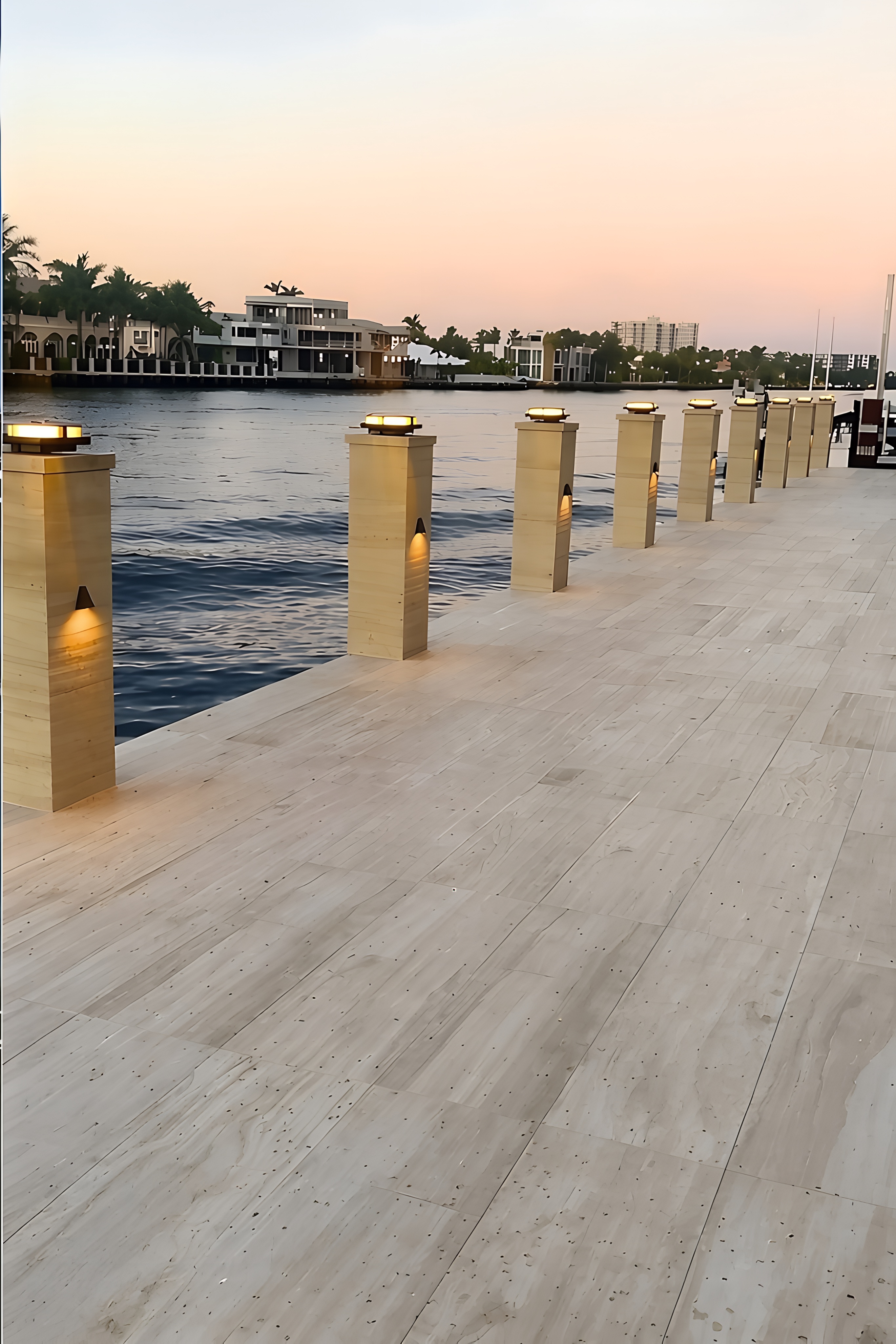 Illuminated pillar lights along a stone-paved waterfront walkway beside a canal with residential homes at dusk.