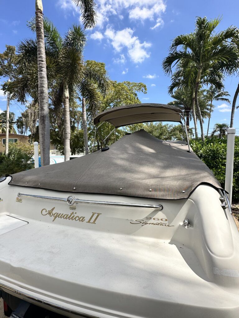 Stern of a white Aquatica II 260 Signature boat with a gray canvas cover and bimini top, parked among palm trees.
