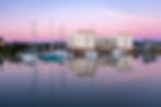 Moored boats and a waterfront building reflected in calm water at dusk at a marina.