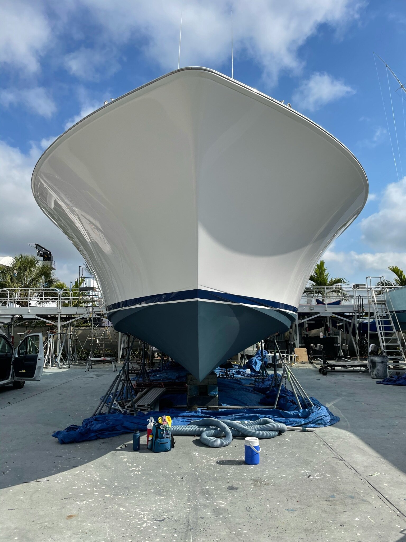 Large white sportfishing boat with freshly painted blue hull resting on stands at a boatyard, with cleaning supplies and a vacuum hose laid out on blue tarps.