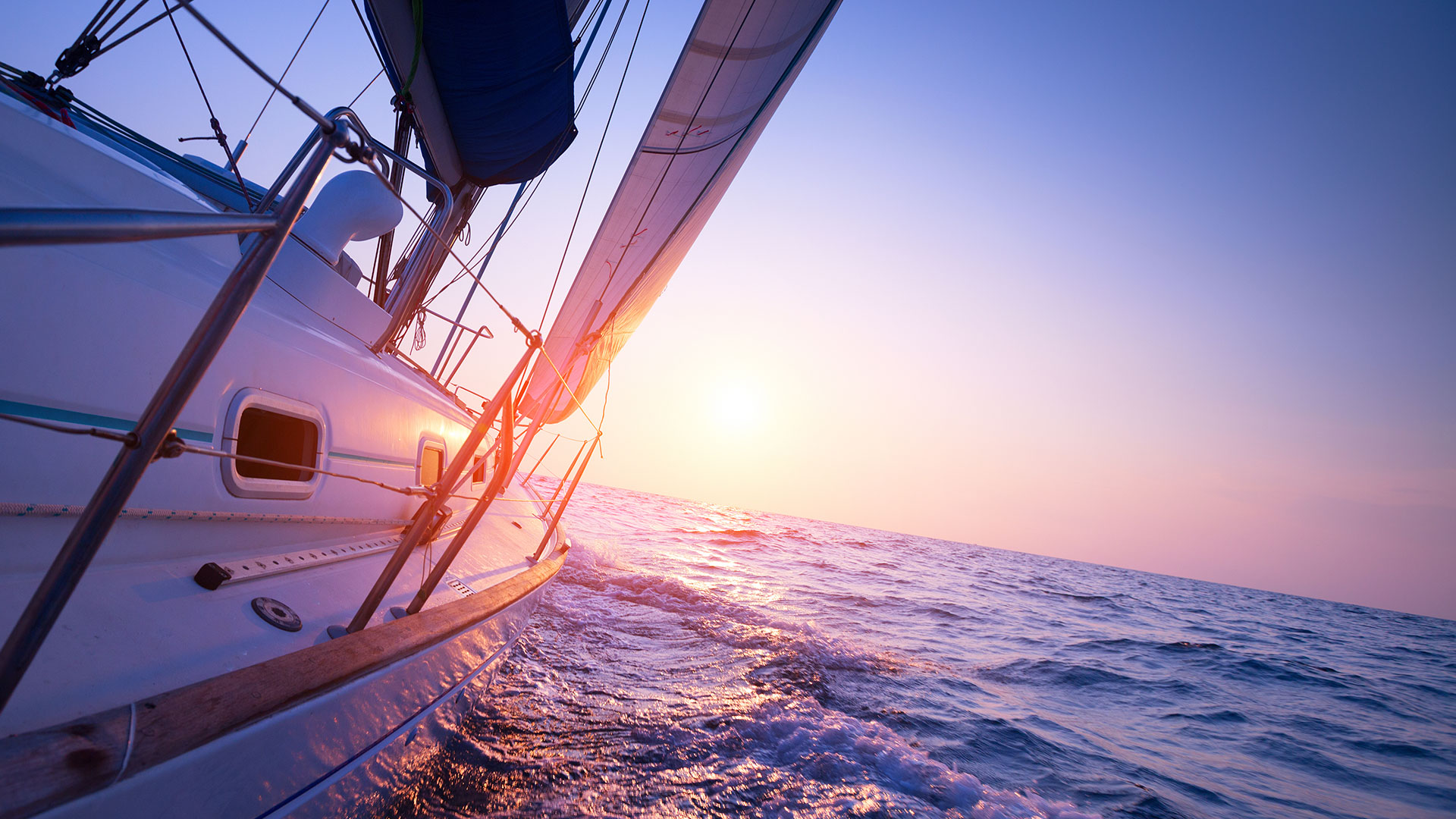 White sailboat with sails trimmed, heeling across open water toward a setting sun.