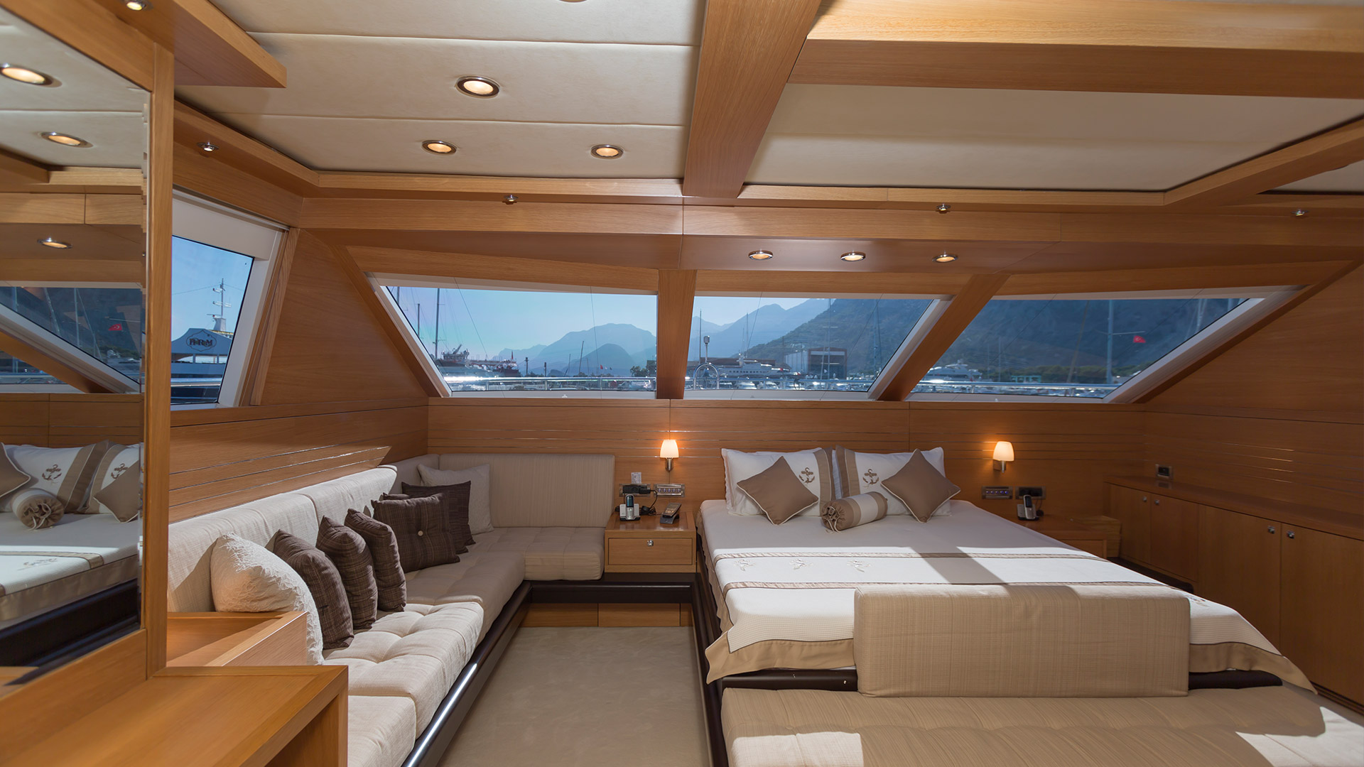 Luxury yacht saloon cabin with wood paneling, cream upholstered seating and a double berth, with a marina and mountains visible through the windows, Turkey.