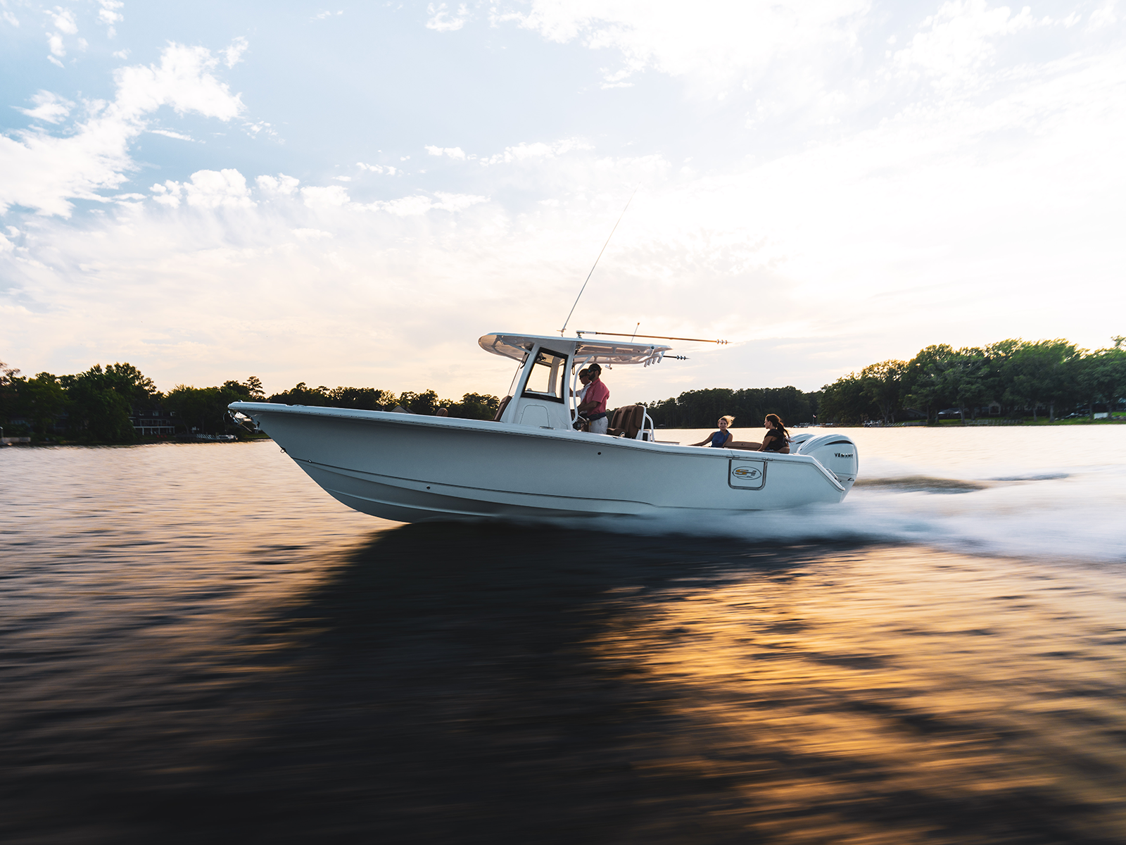 White center-console boat with a T-top and outboard motor cruising across a calm lake at sunset with three passengers aboard.
