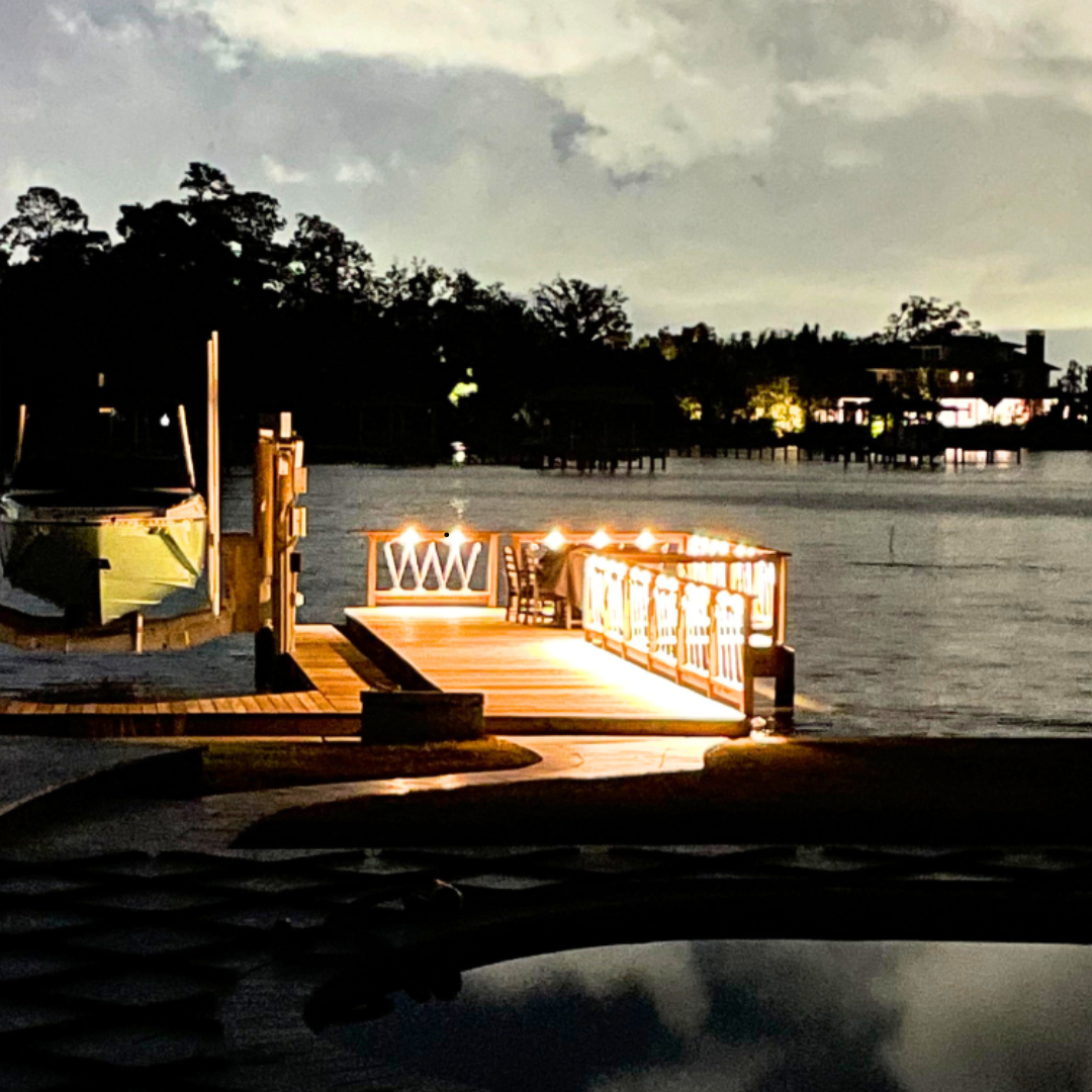 Illuminated wooden dock with string lights and seating overlooking the water at dusk, with a boat on a lift beside it.