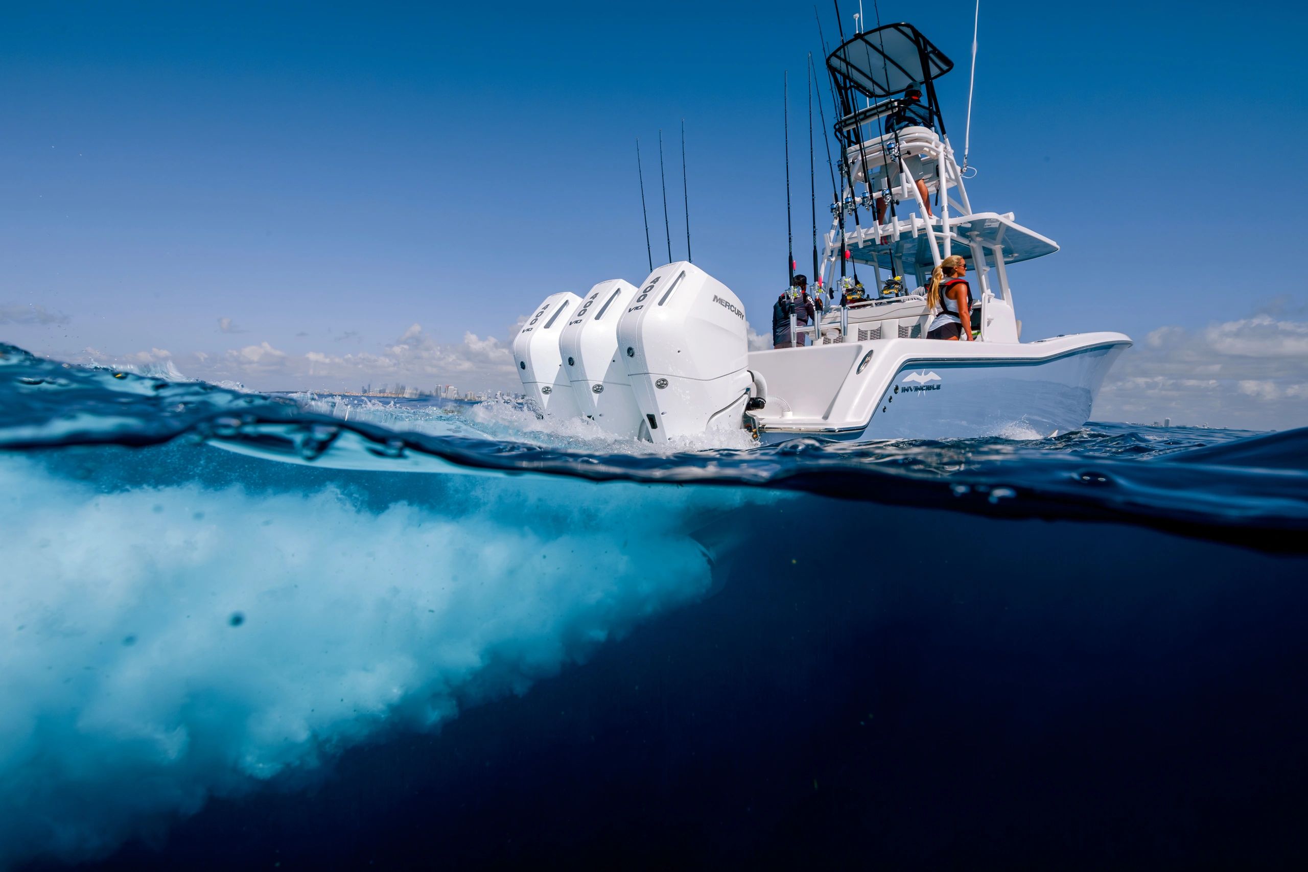Invincible fishing boat powered by three Mercury 400 V10 outboards kicking up spray on open blue water with a city skyline on the horizon.